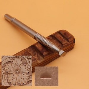 May include: A metal leathercraft tool with a ridged handle and angled blade rests on a wooden holder. The image includes a close-up of a floral leather carving and a sample impression, showcasing the tool's potential for detailed work.