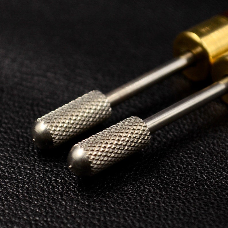 May include: Two silver metal nail drill bits with a diamond-shaped pattern on the surface. The bits are attached to a metal rod and are used for nail care.