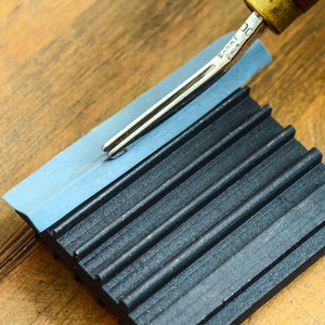 May include: A close-up of a leather stitching tool being used on a black and blue leather working tool. The tool has a wooden handle and a metal blade.