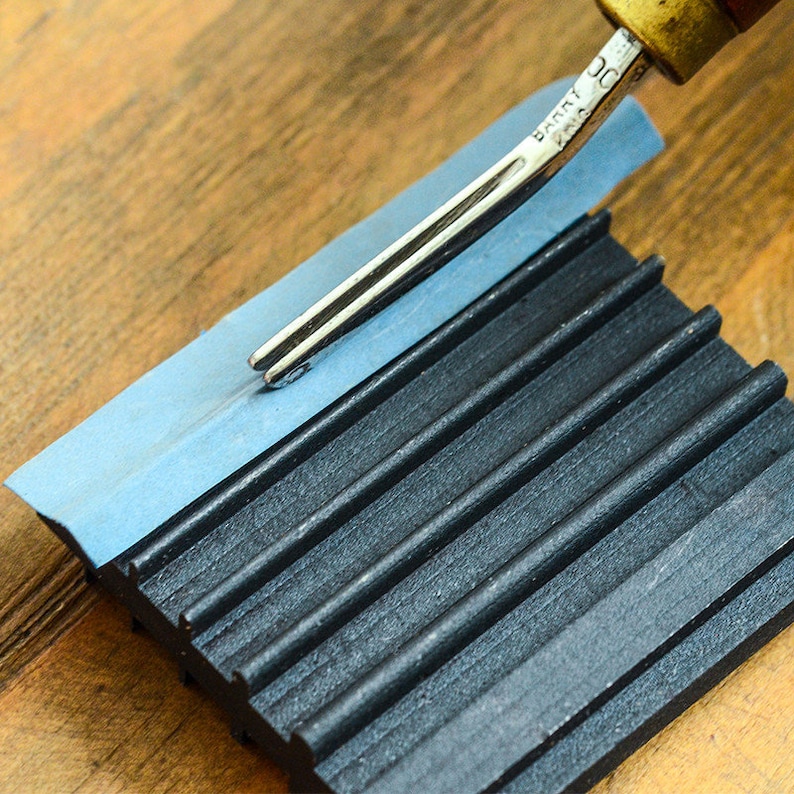 May include: A close-up of a leather stitching tool being used on a black and blue leather working tool. The tool has a wooden handle and a metal blade.