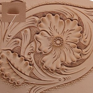 May include: Close-up of a tan leather surface with intricate floral and leaf tooling. The design features a large, detailed flower at the center, surrounded by swirling leaves and decorative patterns. The leather has a natural, textured appearance.