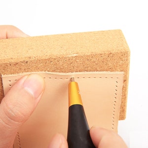 K&ouml;nnte beinhalten: Eine Person verwendet ein Leder-N&auml;hwerkzeug, um L&ouml;cher in ein St&uuml;ck Leder zu stanzen. Das Leder liegt auf einem Korkblock.