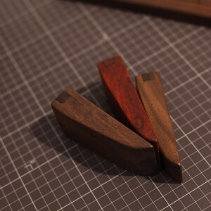May include: Three wooden wedges, one red, one brown, and one dark brown, with metal accents. The wedges are arranged in a triangular shape on a gray grid surface.