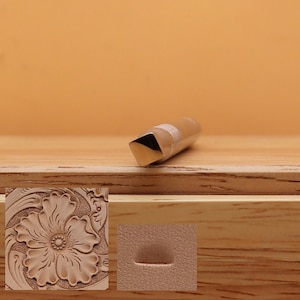 May include: A metal leathercraft tool with a square end and a cylindrical body. The tool is designed for creating decorative patterns on leather. The image also shows a sample of a floral design and a curved line created with the tool.