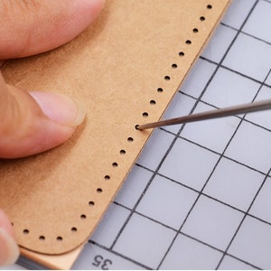May include: A close-up of a brown leather piece with a row of evenly spaced holes along the edge. A needle is being used to punch through the holes.