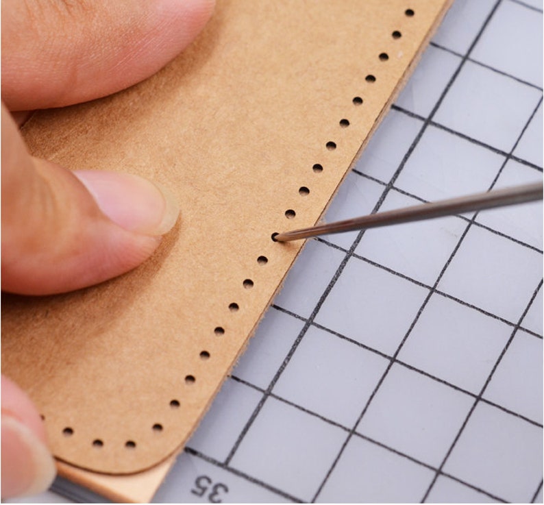 May include: A close-up of a brown leather piece with a row of evenly spaced holes along the edge. A needle is being used to punch through the holes.
