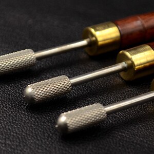 May include: Three leather working tools with wooden handles and metal tips. The tools have a brass colored metal band at the top of the handle.