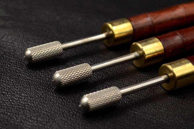 May include: Three leather working tools with wooden handles and metal tips. The tools have a brass colored metal band at the top of the handle.
