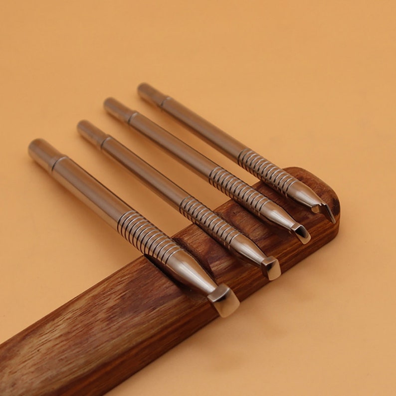 May include: Four silver metal tools with different shaped tips, resting on a dark wooden holder. The tools have a cylindrical body with a ridged section near the tip. The background is a solid light brown color.