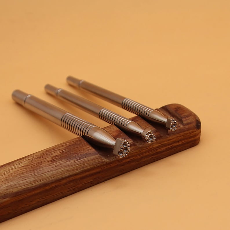 Op de afbeelding: Drie zilveren metalen stempels met verschillende ontwerpen worden getoond in een houten houder. De stempels hebben een getextureerde, geribbelde handgreep en worden gebruikt voor knutselen en leerbewerking.