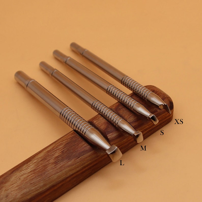 May include: Four stainless steel leathercraft tools with different tip shapes, labeled XS, S, M, and L, are displayed on a wooden holder. The tools have a cylindrical body with a textured grip area. The background is a warm, neutral tone.