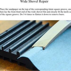 May include: A close-up of a blue sanding block being used to smooth the edge of a metal shovel. The shovel is resting on a black grooved surface.
