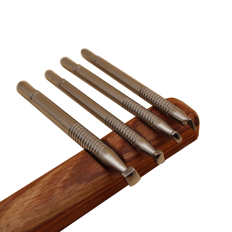 May include: Set of four stainless steel tools with various shaped tips, resting on a dark wooden holder. The tools have a metallic finish and are designed for detailed work. The wooden holder provides a contrasting warm tone.