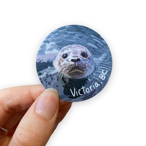 Puede incluir: Una pegatina redonda con una ilustraci&oacute;n de dibujos animados de la cara de una foca asomando la cabeza fuera del agua. La foca tiene un pelaje gris claro y blanco y ojos negros. La pegatina tiene el texto "Victoria, BC" y "Meaghan Crow" en letras blancas.