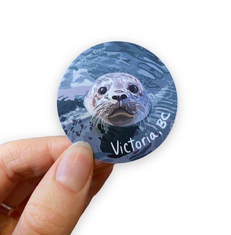 Puede incluir: Una pegatina redonda con una ilustraci&oacute;n de dibujos animados de la cara de una foca asomando la cabeza fuera del agua. La foca tiene un pelaje gris claro y blanco y ojos negros. La pegatina tiene el texto "Victoria, BC" y "Meaghan Crow" en letras blancas.