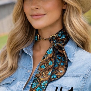May include: A woman wearing a straw cowboy hat and a denim shirt. A patterned scarf with blue floral and paisley designs is around the neck, secured with a gold-colored clasp. The number 1 is printed in black.