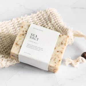 Puede incluir: Una barra de jabón blanca con una etiqueta que dice "BRAND NAME SEA SALT SOAP BAR" y "Jabón natural hecho a mano formulado para nutrir profundamente e hidratar tu piel 4 oz (113g)". El jabón está en una bolsa de malla blanca con una cuenta de madera.