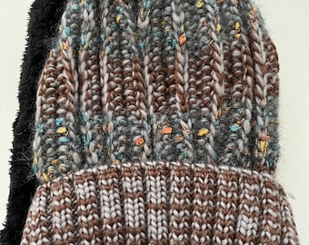 May include: A brown, gray, and blue knit beanie with a fluffy gray pom pom on top. The beanie has a brown and gray knit cuff and a black faux fur ear flap.