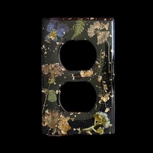 May include: Black electrical outlet cover with embedded dried flowers and gold leaf accents. The cover features two circular openings for the outlets and is surrounded by a black border. The flowers include purple, white, and green varieties.