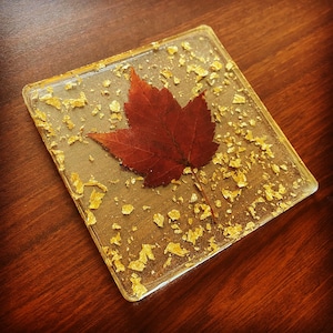 May include: A square clear resin coaster with a red maple leaf pressed inside. The coaster is covered in gold glitter.