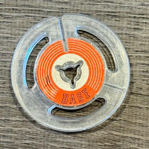 May include: A circular clear plastic reel featuring an orange center label with white concentric rings and the word BASE printed in bold orange capital letters. The center of the reel has a three-pronged cutout design for mounting purposes.