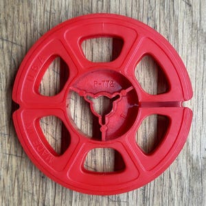 May include: A bright red film reel with a circular design and open spokes. The reel has the text "USA" and "MADE IN" imprinted on it. The center of the reel has the text "P-772".