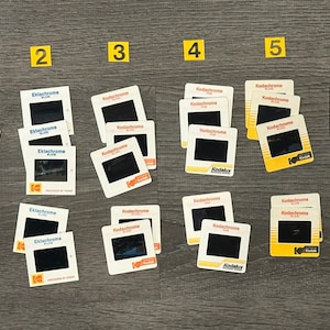 May include: A collection of vintage Kodachrome and Ektachrome film slides, arranged on a dark wooden surface. The slides are in white and yellow frames, with black film areas. Numbers 1 to 6 are on yellow squares.