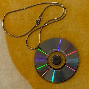 May include: A silver necklace featuring a repurposed CD pendant. The CD has a reflective surface with rainbow hues and a central metallic component. The necklace chain is thin and delicate, with a simple clasp.
