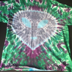 May include: A tie-dye t-shirt with a swirling pattern of green, purple, and blue. The center of the shirt features a faded gray alien face.