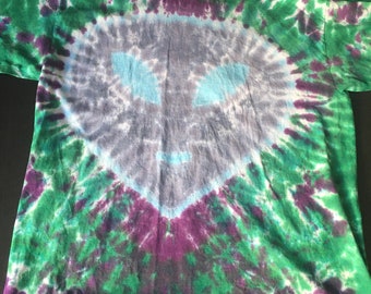 Puede incluir: Una camiseta tie-dye con un patrón de remolino de verde, morado y azul. El centro de la camiseta presenta una cara de extraterrestre gris descolorida.