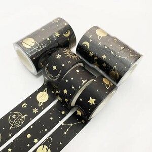 May include: Five rolls of black and gold washi tape with celestial designs. The tape features gold stars, moons, planets, and constellations on a black background.