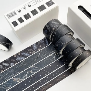 May include: Set of six black and gold washi tapes with different patterns, including marble, stars, and geometric designs. The tapes are rolled up and arranged in a line.