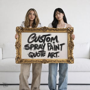 以下が含まれることがあります： 黒いスプレーペイントで「CUSTOM SPRAY PAINT QUOTE ART」と書かれた額入りのアートワーク。フレームは金色の装飾が施されています。2人の人が白い背景の前でアートワークを持っています。
