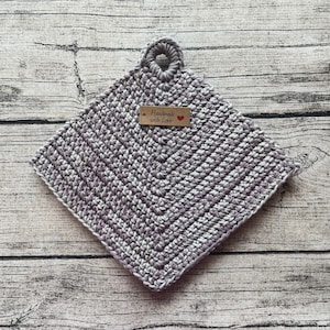 Op de afbeelding: Een gehaakte ovenwant in grijstinten en wit met een lusvormige handgreep. Een houten label met de tekst "Handmade with Love" en een rood hart is bevestigd aan de ovenwant.