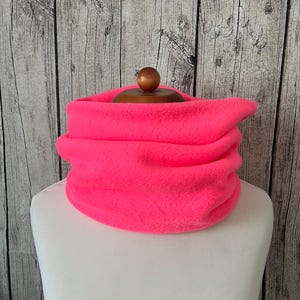 Puede incluir: Un calentador de cuello de forro polar rosa neón vibrante, diseñado para mantener el cuello caliente. El calentador de cuello se muestra en un busto de maniquí blanco sobre un fondo de madera rústica. El material de forro polar es suave y parece estar doblado sobre sí mismo.