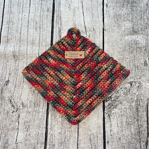 Könnte beinhalten: Ein handgefertigter, quadratischer Topflappen aus Häkelarbeit mit Zickzackmuster. Er hat eine Schlaufe zum Aufhängen und ein kleines Etikett mit der Aufschrift "Handmade with Love". Die Farbpalette umfasst Rot, Orange, Grün und Braun.