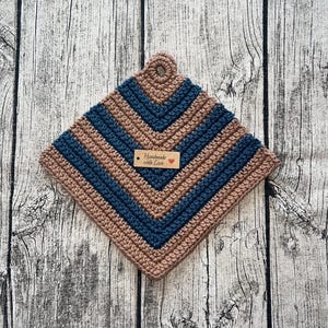Könnte beinhalten: Ein handgefertigter, quadratischer Topflappen aus Häkelarbeit in den Farbtönen Beige und Marineblau. Das Design zeigt abwechselnde Streifen und ein kleines Etikett mit der Aufschrift "Handmade with Love". Der Topflappen hat eine Schlaufe zum Aufhängen.