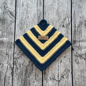Op de afbeelding: Een vierkante gehaakte pannenlap in marineblauw en geel, met een ophanglus. Het ontwerp heeft chevronstrepen. Een klein houten label leest "Handmade with Love" met een hartsymbool. Ontworpen voor gebruik in de keuken.