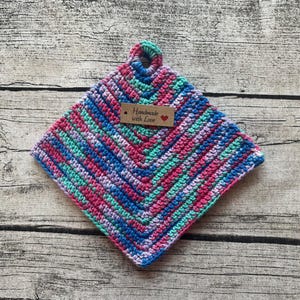 Könnte beinhalten: Ein bunter gehäkelter Topflappen in den Farben Blau, Rosa, Grün und Lila. Der Topflappen hat einen Schlaufe zum Aufhängen und ein braunes Etikett mit der Aufschrift "Handmade with Love" und einem roten Herz.