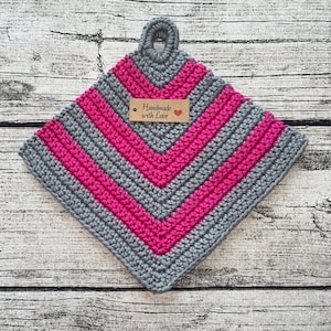 Könnte beinhalten: Grauer und pinkfarbener gehäkelter Topflappen mit einer Schlaufe. Der Topflappen hat ein braunes Etikett mit der Aufschrift "Handmade with Love" und einem roten Herzen.