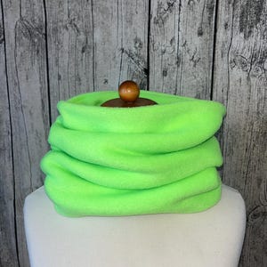 Może przedstawiać: Neonowo-zielony ocieplacz na szyję z polaru. Ocieplacz na szyję jest złożony na białej głowie manekina. Drewniana gałka jest widoczna na górze ocieplacza. Tło to szara drewniana ściana.