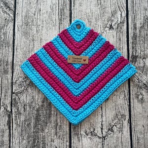 Op de afbeelding: Een vierkante, handgemaakte gehaakte pannenlap in heldere kleuren. Het ontwerp heeft afwisselende rijen turquoise en magenta, met een klein label met de tekst "Handmade with Love". Er is een lus aan de bovenkant om op te hangen.