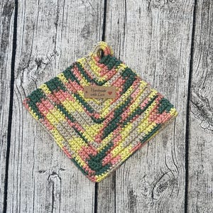 Peut inclure: Un dessous de plat crocheté dans des tons de vert, jaune, rose et beige avec une étiquette brune indiquant "Handmade with Love".