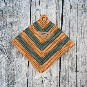 Könnte beinhalten: Ein gehäkelter Topflappen mit einem grün-orangen Streifenmuster. Er hat eine Schlaufe oben und ein braunes Etikett mit der Aufschrift "Handmade with Love".