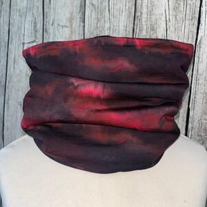 Op de afbeelding: Een tie-dye nekwarmer in rood en zwart, weergegeven op een mannequin. De stof is geplooid, waardoor een gelaagd effect ontstaat. Dit accessoire kan worden gedragen als gezichtsbedekking of nekwarmer.