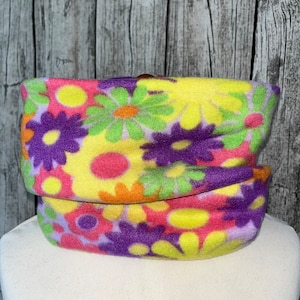 Könnte beinhalten: Ein farbenfroher Fleece-Halswärmer mit einem Blumenmuster in den Farben Gelb, Rosa, Lila, Grün und Orange. Die Blumen sind stilisiert und ähneln Gänseblümchen.