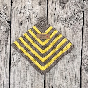 Op de afbeelding: Een grijze en gele gestreepte gehaakte ovenwant met een lus om op te hangen. De ovenwant heeft een bruin label met de tekst "Handmade with Love".