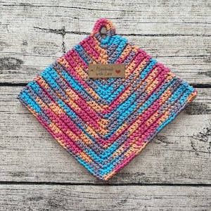 Peut inclure: Un dessous de plat carré fait à la main au crochet, avec un motif en chevrons. Le dessous de plat est doté d'une boucle pour l'accrocher et est réalisé avec des fils roses, bleus, oranges et violets. Une petite étiquette indique "Handmade with Love".