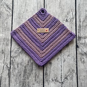 Op de afbeelding: Een handgemaakte, vierkante gehaakte pannenlap in tinten paars en mauve. Het ontwerp heeft een chevronpatroon en een klein label met de tekst "Handmade with Love" met een hartsymbool. Er zit een lus aan de bovenkant om op te hangen.
