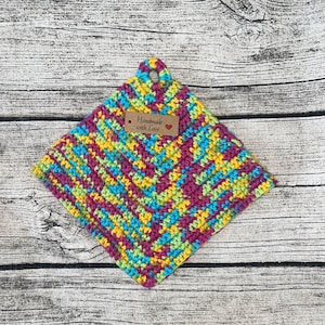 Może przedstawiać: Kolorowy, szydełkowy uchwyt na garnek z brązową etykietą z napisem "Handmade with Love" i czerwonym sercem.